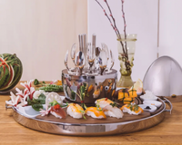 The Christofle MOOD Asia Tray - Stainless Steel displays assorted sushi and garnishes on a wooden table, with a metal bowl of silverware at the center, complemented by decorative accents in the background.