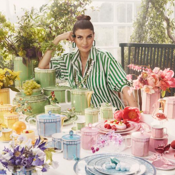 A woman in a green-and-white striped shirt sits at a colorful table set with the Swarovski x Rosenthal Signum Tea Pot Small, pastel cups and plates, fruits, and flowers, surrounded by lush greenery in a bright, sunlit room.