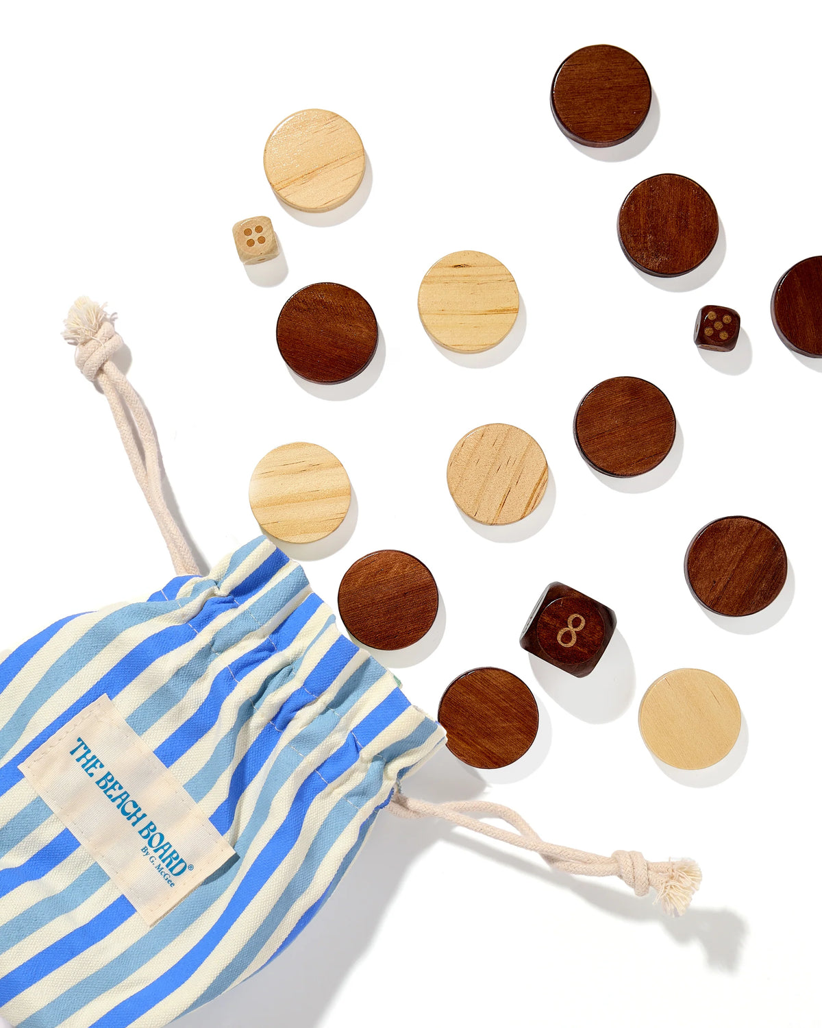 A blue and white striped drawstring bag labeled “Southampton Beach Backgammon” by G. McGee spills round wooden game pieces and two small wooden dice onto a white surface, inspired by a luxury beach towel.