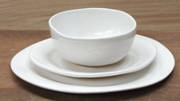 The Urban Dinnerware Matte White set by Alex Marshall Studios features handmade ceramic plates with a bowl stacked on a salad and dinner plate, arranged on wood with a matching wooden background.