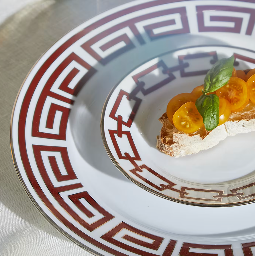 A slice of bread with white cheese, yellow cherry tomatoes, and basil rests on a Catene Scarlatto dinner plate by Ginori 1735, featuring a red and gold geometric border, atop a light tablecloth.