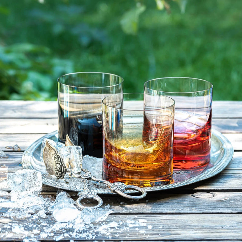Three colorful drinks in Moser’s Whiskey Set DOF Glass 370 ml tumblers sit on a silver tray with vintage tongs and melting ice, arranged on an outdoor wooden table with a softly blurred green background.