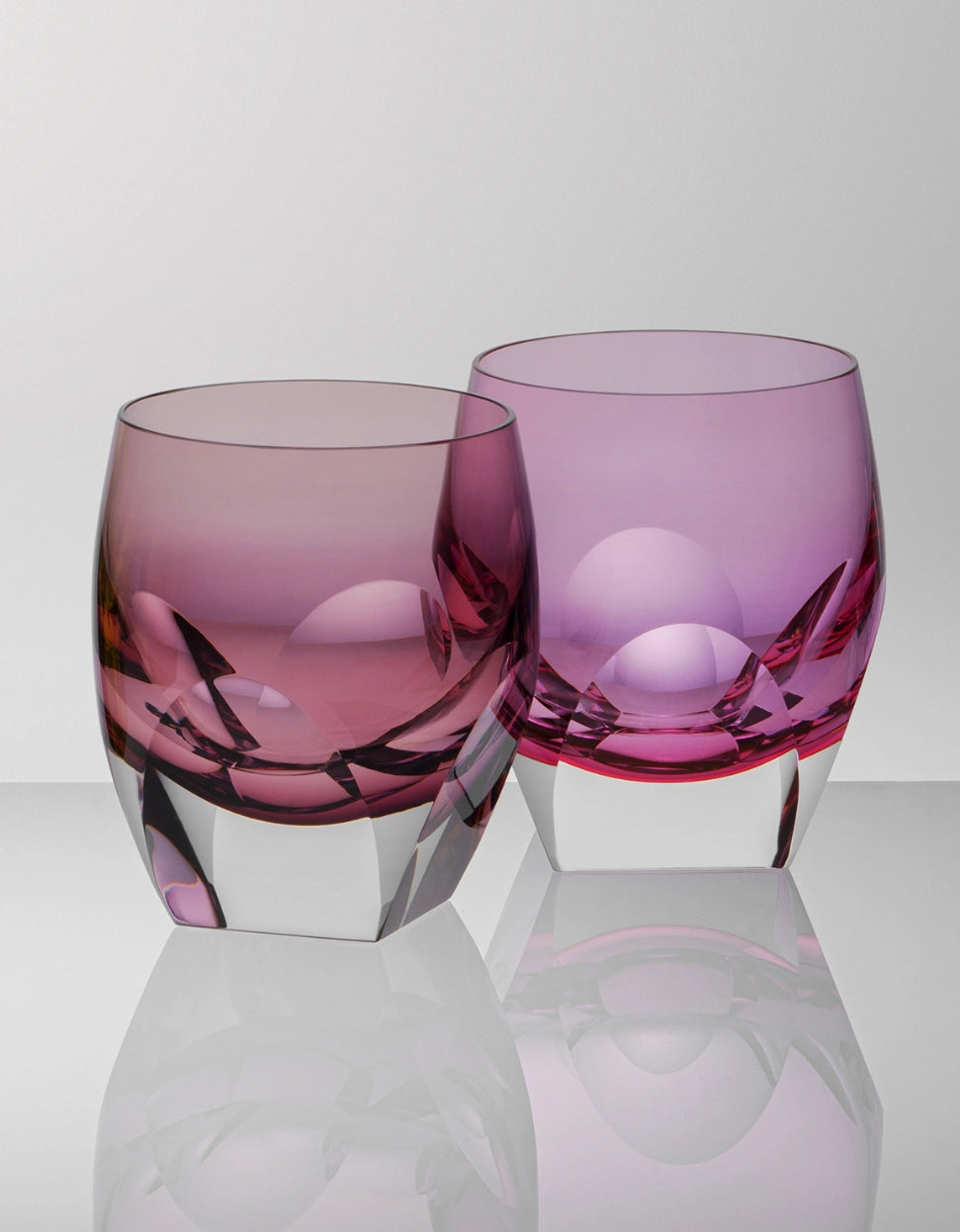 The Moser Bar Spirit Tumbler Shot Glass 45ml, featuring thick clear bases and pink and purple glass, sits side by side on a reflective surface—an elegant addition to any bar collection.