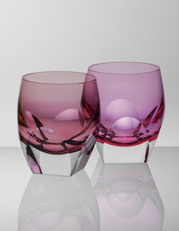 The Moser Bar Spirit Tumbler Shot Glass 45ml, featuring thick clear bases and pink and purple glass, sits side by side on a reflective surface—an elegant addition to any bar collection.