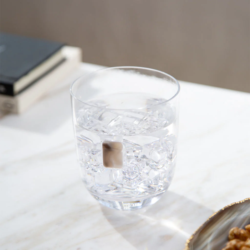 An Elevo DOF Glass by Anna New York in smoke agate, filled with ice water, rests on a white marble surface. A book and another object are blurred in the background, creating a serene setting. Set includes 2 glasses.