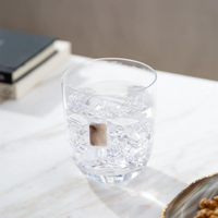 An Elevo DOF Glass by Anna New York in smoke agate, filled with ice water, rests on a white marble surface. A book and another object are blurred in the background, creating a serene setting. Set includes 2 glasses.