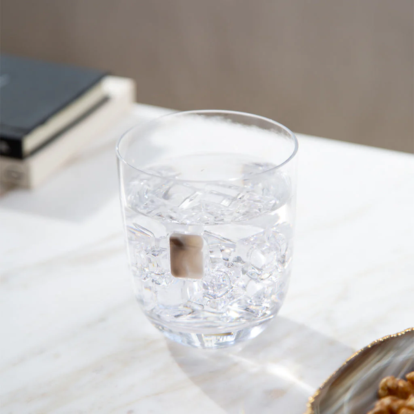 An Elevo DOF Glass by Anna New York in smoke agate, filled with ice water, rests on a white marble surface. A book and another object are blurred in the background, creating a serene setting. Set includes 2 glasses.
