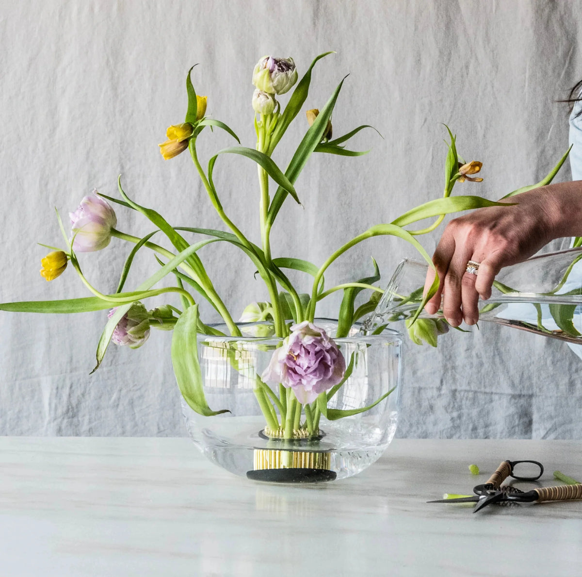 A person waters purple and yellow tulips arranged in a handmade Burlington Bowl by Simon Pearce on a white surface, with gardening shears and twine nearby.