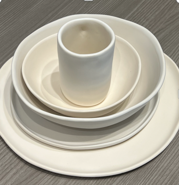 A set of four Urban Dinnerware Matte White plates by Alex Marshall Studios, handmade in varying sizes and stacked with a small cup on top, shown on a wood-textured gray surface.