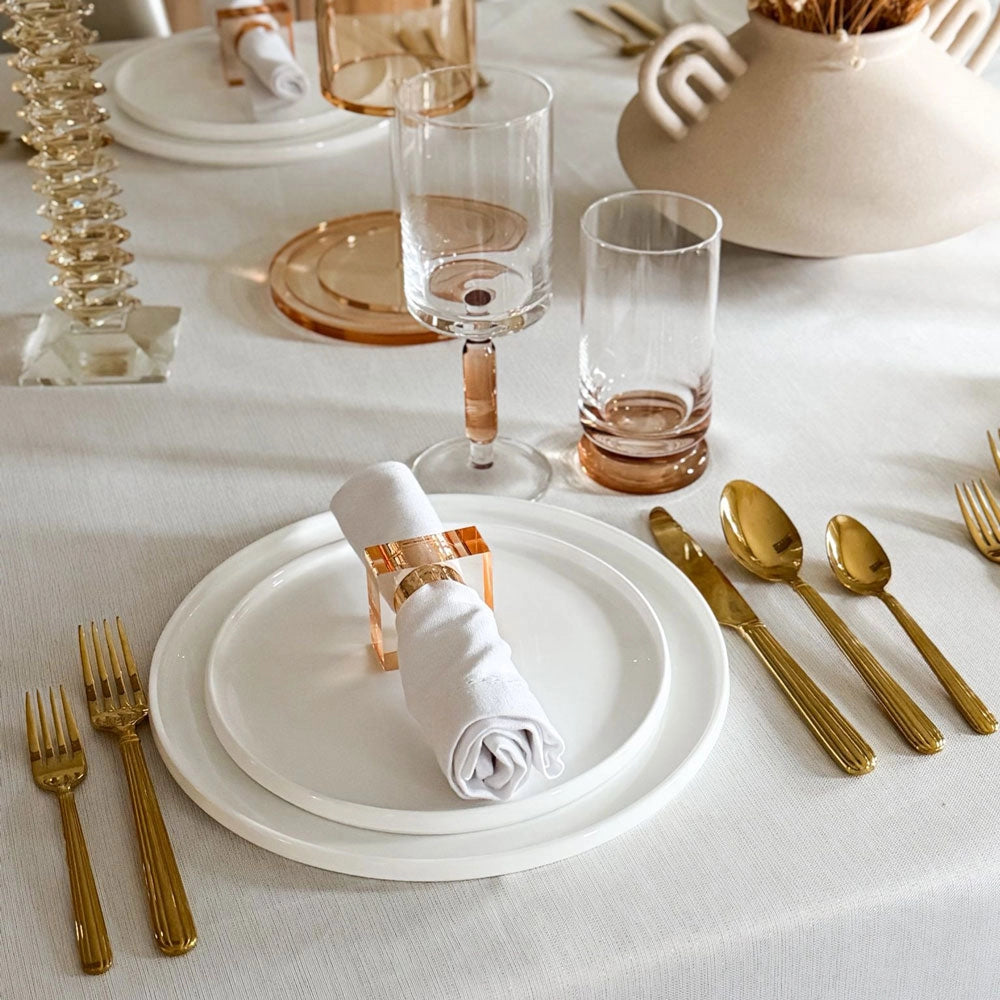 A chic table setting featuring white plates, a rolled napkin held by the Lucite Cube Napkin Ring Set of 4 - Amber from BT- Shalom, gold cutlery, clear and pink glasses, all displayed on a white tablecloth with stylish decorative accents in the background—an ideal showcase of contemporary table decor.