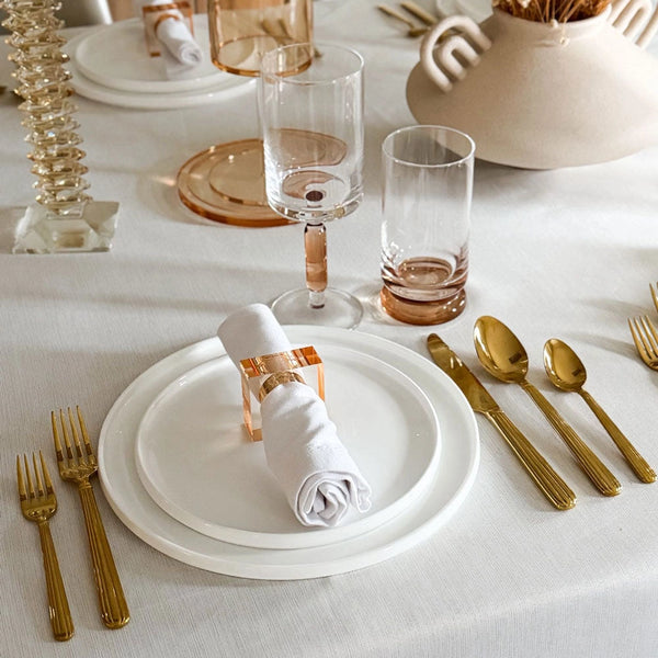 A chic table setting featuring white plates, a rolled napkin held by the Lucite Cube Napkin Ring Set of 4 - Amber from BT- Shalom, gold cutlery, clear and pink glasses, all displayed on a white tablecloth with stylish decorative accents in the background—an ideal showcase of contemporary table decor.