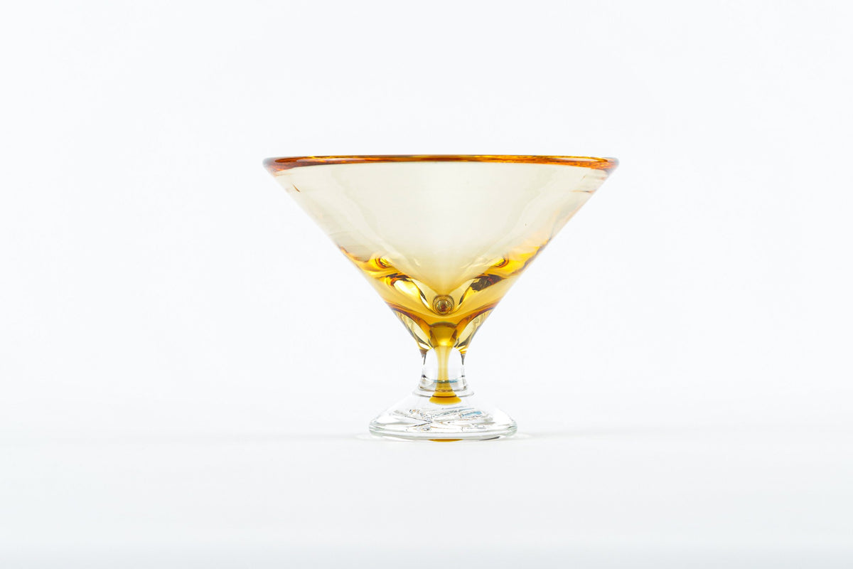 The Footed Martini Glass by Tyler Barry Design features hand-blown glass with a golden rim, partially filled with amber liquid and displayed on a plain white background—an elegant example of modern barware.