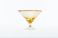 The Footed Martini Glass by Tyler Barry Design features hand-blown glass with a golden rim, partially filled with amber liquid and displayed on a plain white background—an elegant example of modern barware.