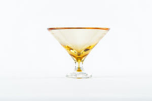 The Footed Martini Glass by Tyler Barry Design features hand-blown glass with a golden rim, partially filled with amber liquid and displayed on a plain white background—an elegant example of modern barware.