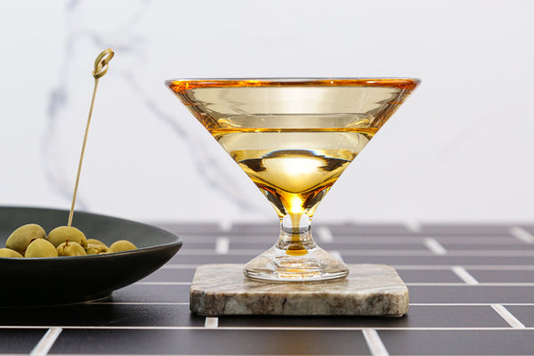 A cocktail in a wide, yellow-tinted Footed Martini Glass by Tyler Barry Design sits on a marble coaster beside green olives with a pick, all atop black tiles—a modern barware statement.