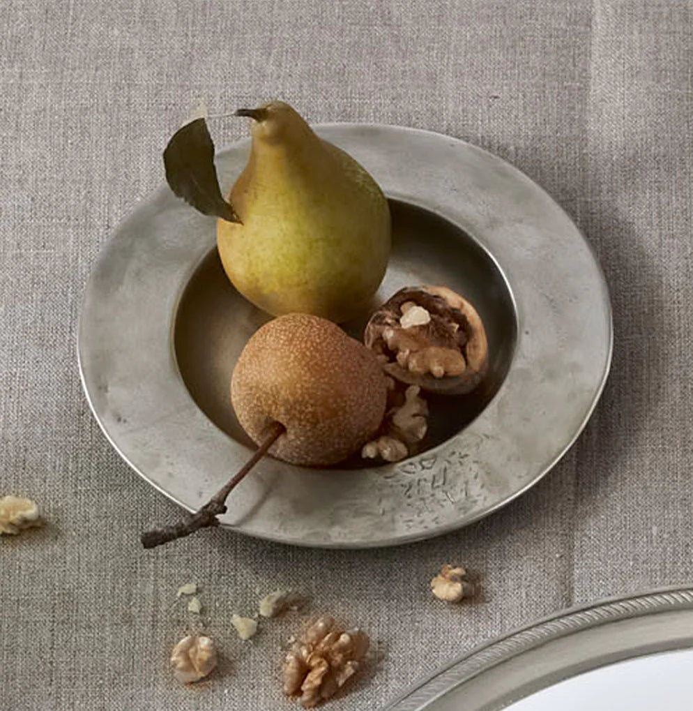 The MATCH Antique Bottle Coaster by MATCH, made of lead-free handmade Italian pewter, holds a small brown pear with a stem, a cracked walnut, and walnut pieces. More walnut pieces are scattered on the beige tablecloth beside it.