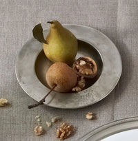 The MATCH Antique Bottle Coaster by MATCH, made of lead-free handmade Italian pewter, holds a small brown pear with a stem, a cracked walnut, and walnut pieces. More walnut pieces are scattered on the beige tablecloth beside it.