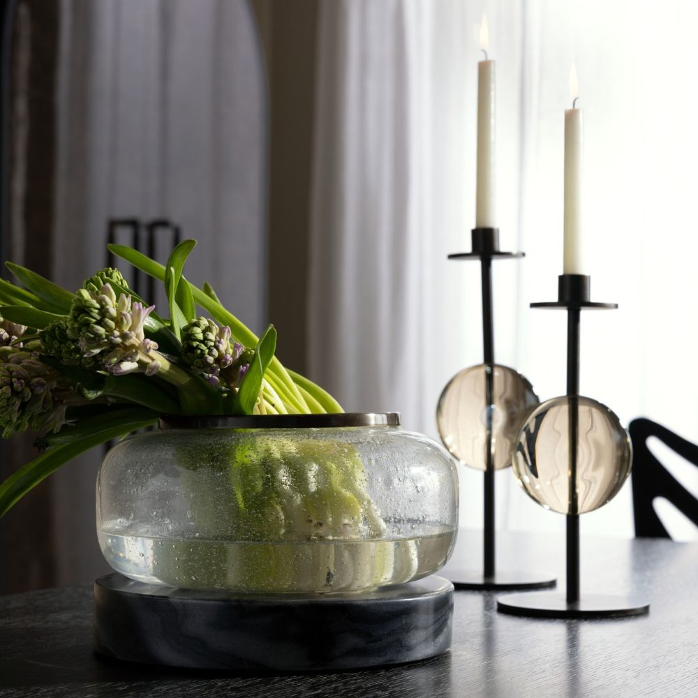 A glass vase with green hyacinths sits on a dark table next to the Terrell Candleholders Set of 2 by Arteriors—one candleholder features sleek lines and champagne crystal details, while the other is crafted from bronze-finished steel. Both are topped with white candles and accented by round glass elements, as light filters through sheer curtains in the background.