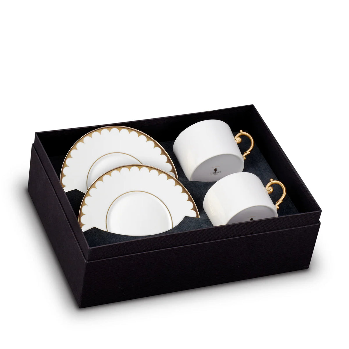 Two L’Objet Aegean Filet Gold Dinnerware teacups with gold handles and two matching scalloped saucers with gold rims, presented in a black gift box.