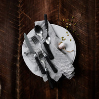 A white plate with a gray striped napkin holds the Vietri Albero Flatware 5 Piece Setting in Elm atop a dark wooden surface, accented by a garlic bulb and scattered green seeds.
