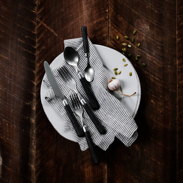 A white plate with a gray striped napkin holds the Vietri Albero Flatware 5 Piece Setting in Elm atop a dark wooden surface, accented by a garlic bulb and scattered green seeds.