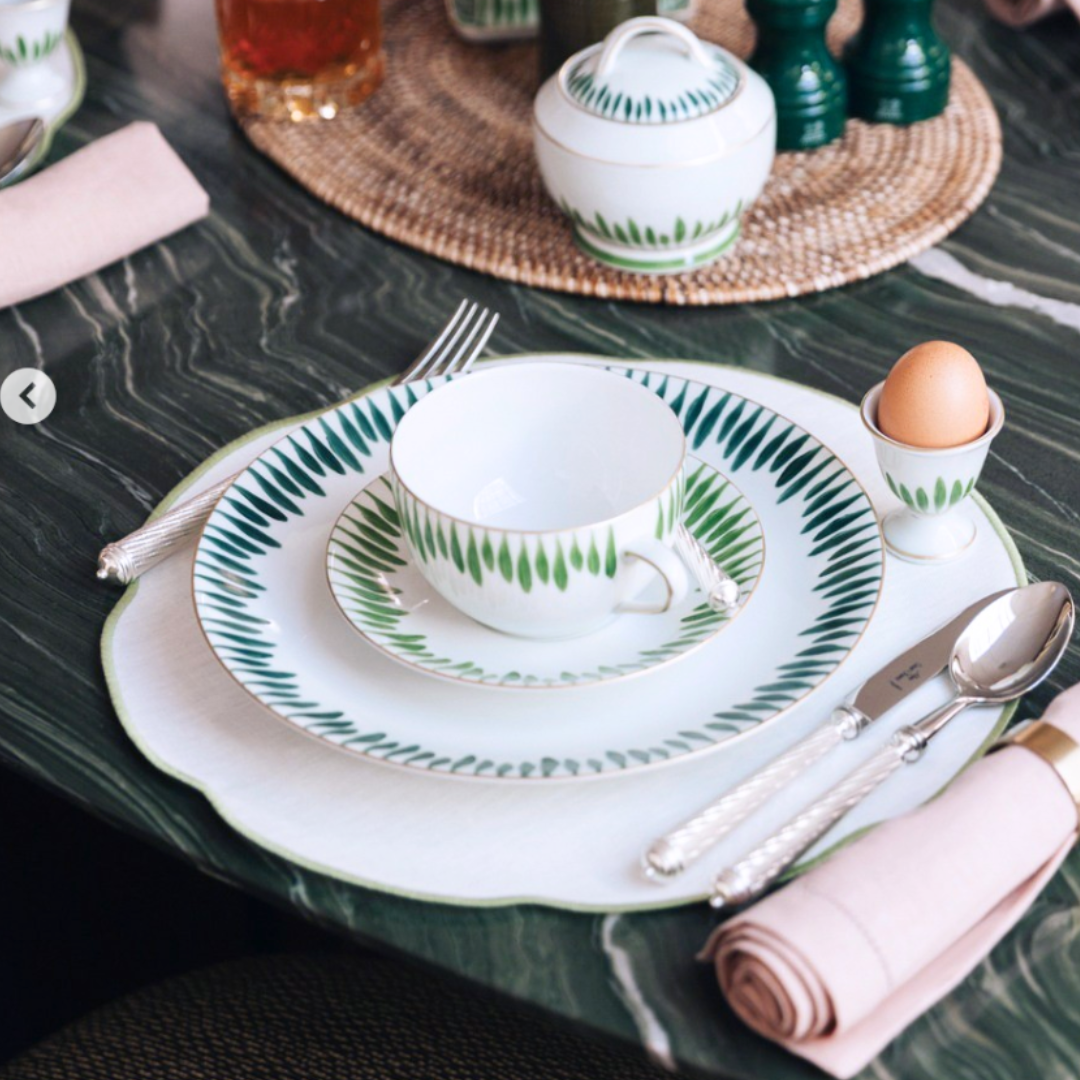 A table setting features Marie Daâge Allee de Cypres 5 porcelain by Marie Daâge—a green and white patterned plate, cup, and saucer—alongside silver flatware, a pink napkin, and a soft-boiled egg in an egg cup on a green marble table.