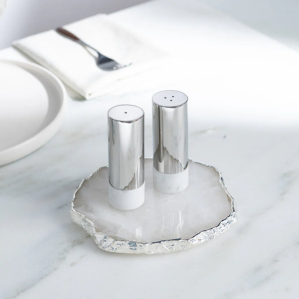 The Anna New York Coluna Dual Salt & Pepper Shakers Set of 2 in marble and silver sits on a round white marble tray with a textured silver edge, adding a modern touch to your dining table or countertop.