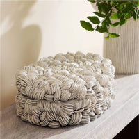 The Arteriors Hampton Box - New Addition, featuring a textured woven-look and lid, sits on a light wood surface beside a ribbed vase with green branches, adding a sculptural accent to your space.