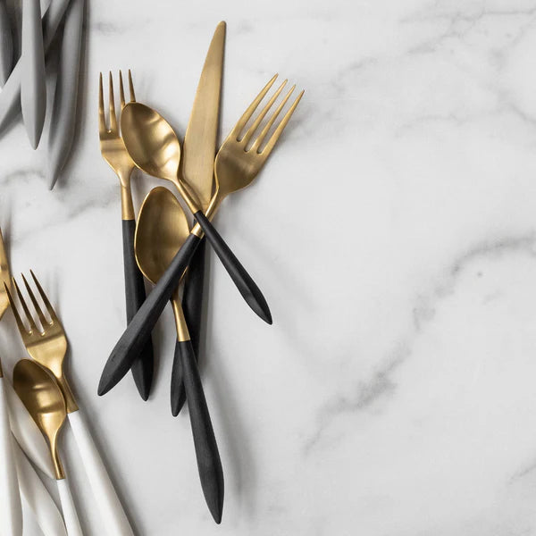 The Vietri Ares Flatware 5 Piece Setting-Black & Gold features brushed gold forks, knives, and spoons with black handles, arranged on white marble, alongside extra 18/10 stainless steel utensils with white handles.