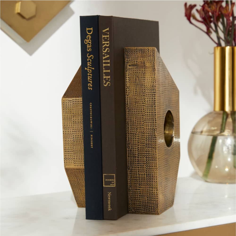 Two books stand upright between the Garrick Bookends Set of 2 by Arteriors, showcasing a geometric antique brass texture on a white surface, with a glass vase of red flowers in the background.