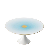 The Aura Dinnerware cake stand by Raynaud is a white porcelain piece, made in France, with blue concentric circles and a gold center on its wide round top, set on a short pedestal base.