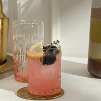 An Aaron Pink Glassware Set of 6 by Blue Pheasant sits in sunlight, with one tumbler filled with a pink drink garnished with lemon, blackberries, and thyme on a gold coaster. A layered dessert is partially visible beside the glasses.