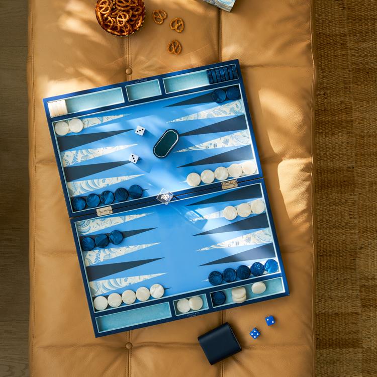 The Addison Ross Palm Beach Backgammon Set – Blue sits on a tan cushioned surface with dice, checkers, and accessories; a few pretzels are scattered nearby.