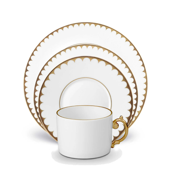 The L'Objet Aegean Filet Gold Dinnerware set includes a porcelain teacup, saucer, and two plates, each featuring scalloped 24k gold rims. The pieces are elegantly stacked with the cup in front of the plates.