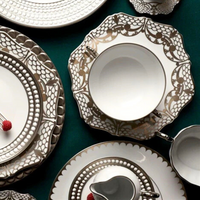 L'Objet Aegean Filet Platinum Dinnerware—plates, bowls, and cups—features elegant white pieces with platinum accents, artfully arranged with silverware on a dark surface to highlight their luxurious design.
