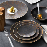 Jaune De Chrome’s Aguirre Dinnerware—a rustic, textured dark set of plates and bowls inspired by luxury tableware—sits arranged with a fork, napkin, wooden salt and pepper shakers, gourmet appetizers, and a silver spoon resting in one bowl.