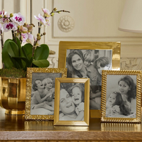 Four black-and-white family photos are displayed on a wooden table in Aerin's Alessio gold frames with a reeded design, next to a potted orchid. The images of smiling adults and children capture happy family moments.