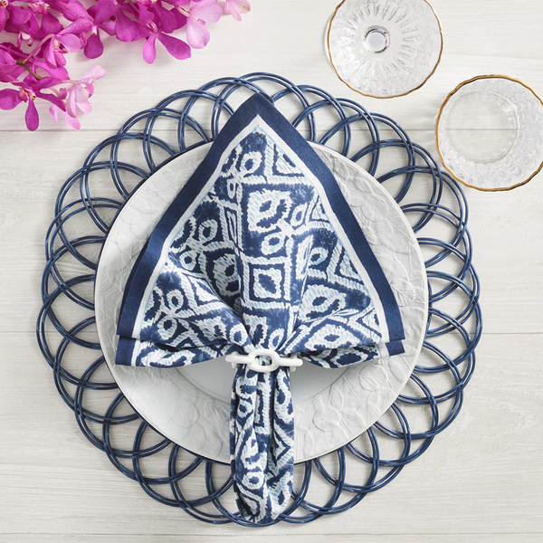 A white plate with a Kim Seybert Alfresco Napkin Navy & Blue Set of 4 rests on a woven blue placemat. Clear glassware and pink flowers are nearby on a light wood table.