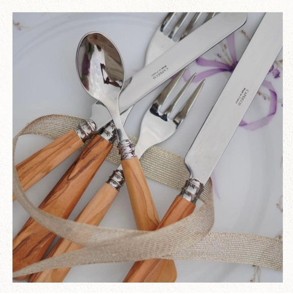 The Capdeco Altea Flatware 5 Piece Setting – Olive Wood features a knife, fork, and spoon with olive wood handles, elegantly arranged on a white plate with a sheer beige ribbon woven through the set.