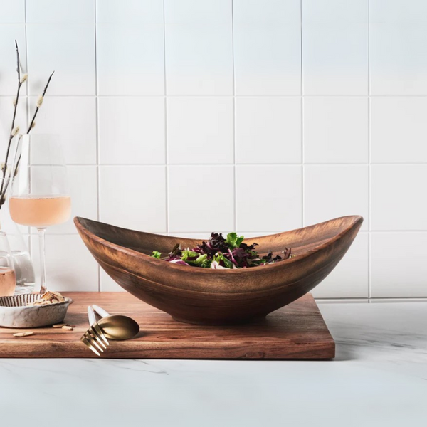 The Andrew Pearce Live Edge Wood Bowl Walnut, handmade in Vermont, holds mixed greens on a wooden board. Gold utensils, a glass of rosé, and a small dish accompany it amid decorative branches against white tile.
