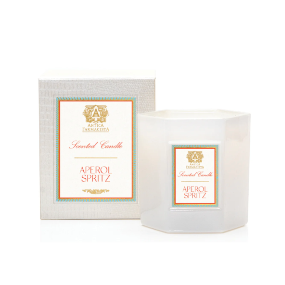 The Antica Farmacista/Harper Aperol Spritz Candle, a white hexagonal citrus-scented candle in a glass holder with gold, orange, and teal accents and logo, sits next to its matching box.