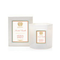 The Antica Farmacista/Harper Aperol Spritz Candle, a white hexagonal citrus-scented candle in a glass holder with gold, orange, and teal accents and logo, sits next to its matching box.