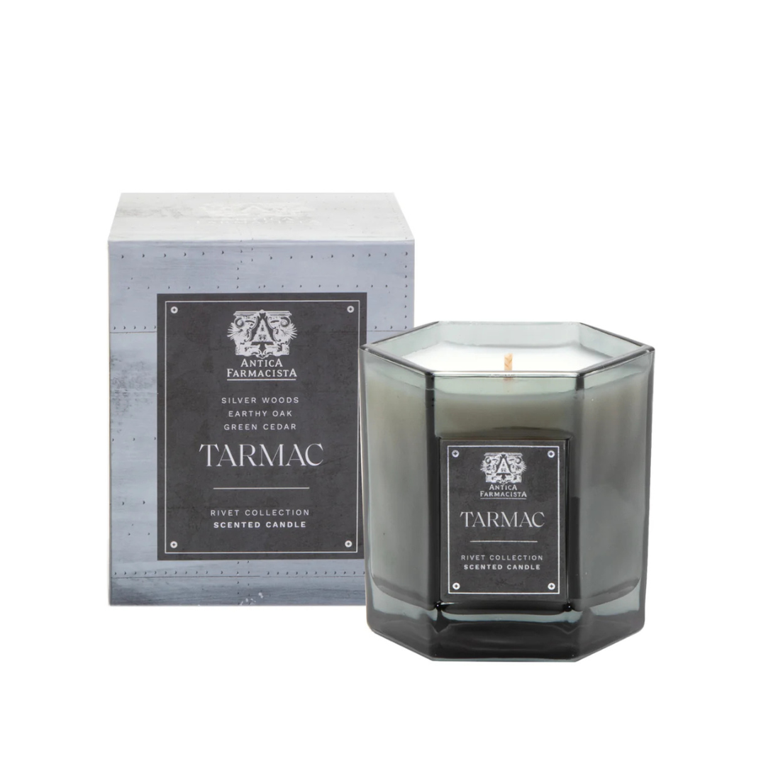 A gray octagonal glass Tarmac Candle by Antica Farmacista/Harper sits before its matching box, both with black and silver labels highlighting scents of silver woods, earthy oak, and green cedar.