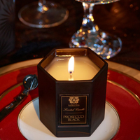 A lit Antica Farmacista/Harper Prosecco Black Candle made from soy-paraffin wax sits on a red and white plate, with a candle snuffer and glassware in the background.