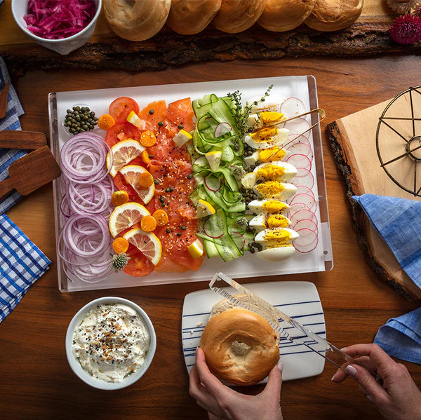 A platter of smoked salmon, veggies, and eggs rests on the Apeloig Solid Acrylic Challah Board & Knife. Someone slices a bagel with the magnetic knife, while cream cheese awaits in a bowl.