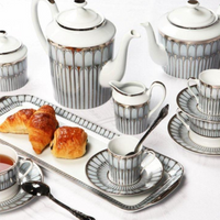 A set of Philippe Deshoulieres BIA Arcades Platinum Dinnerware—porcelain tea/coffee pots, cups, and saucers with a silver-gray geometric pattern—made in France and served with croissants on a matching tray.