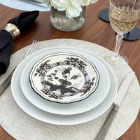 A formal table setting features layered white plates (top with black floral design), silver cutlery, crystal wine glass, black napkin with silver ring—all elegantly arranged on Bodrum’s Tresse Round Placemat Set of 4 atop a wooden table.