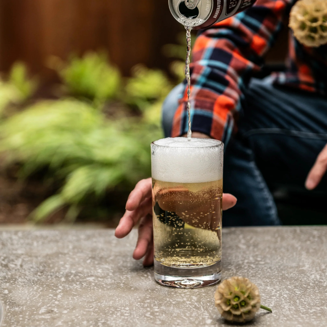 Wearing a plaid shirt, someone pours a fizzy drink from a can into a Simon Pearce Ascutney Tumbler on an outdoor concrete table, with blurred green plants behind—showcasing the timeless elegance of this handblown barware.