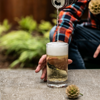 Wearing a plaid shirt, someone pours a fizzy drink from a can into a Simon Pearce Ascutney Tumbler on an outdoor concrete table, with blurred green plants behind—showcasing the timeless elegance of this handblown barware.