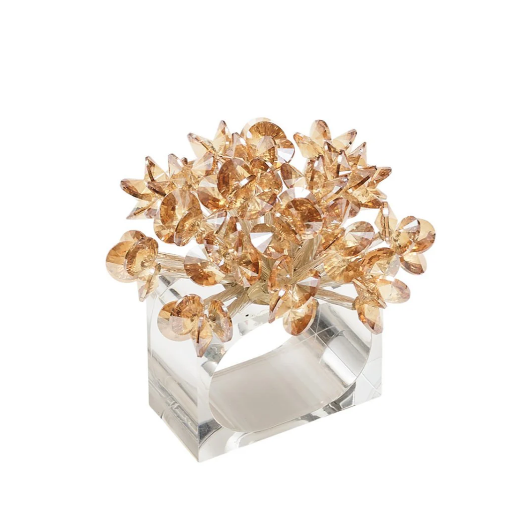 The Kim Seybert Aster Napkin Ring Set of 4 features golden, crystal-like flowers arranged in a bouquet atop a clear, rectangular ring with a cut-out center—adding an elegant touch to your table décor.